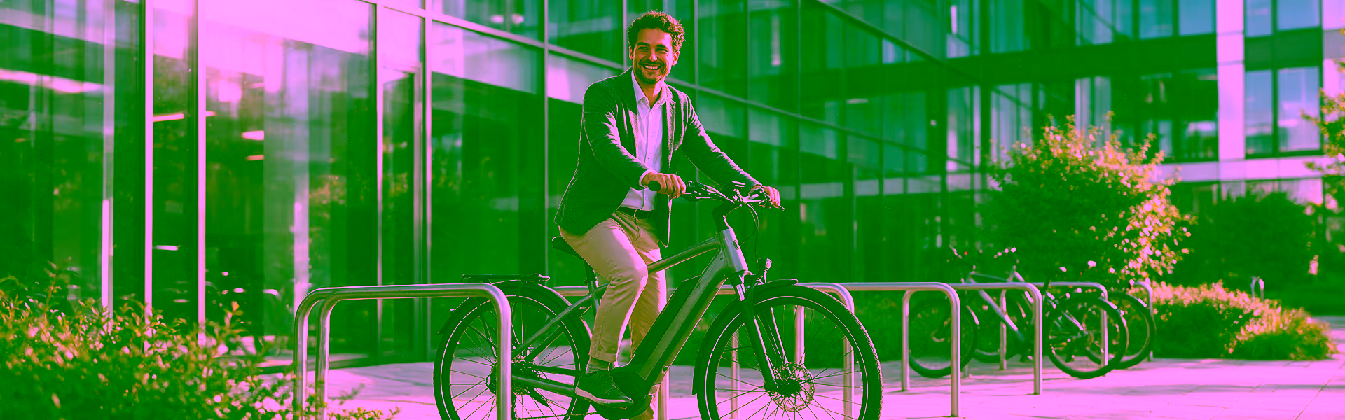 Lächelnder Mitarbeiter im Sakko fährt mit seinem grauen E-Bike an den Fahrradständer vor einem gläsernen Bürogebäude