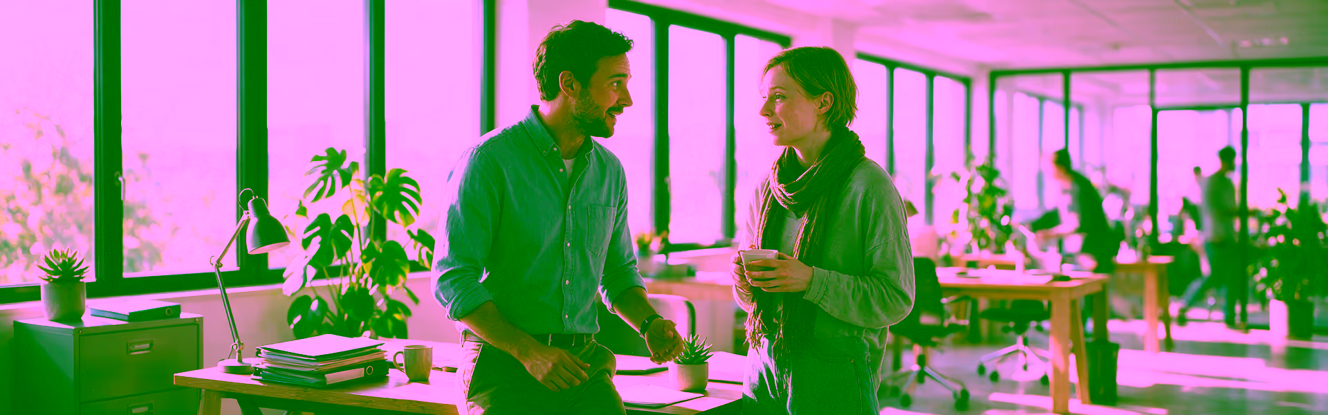 Vorgesetzter spricht im hellen Büro mit einer Mitarbeiterin mit Schal und Teetasse, die trotz Krankschreibung zur Arbeit gekommen ist