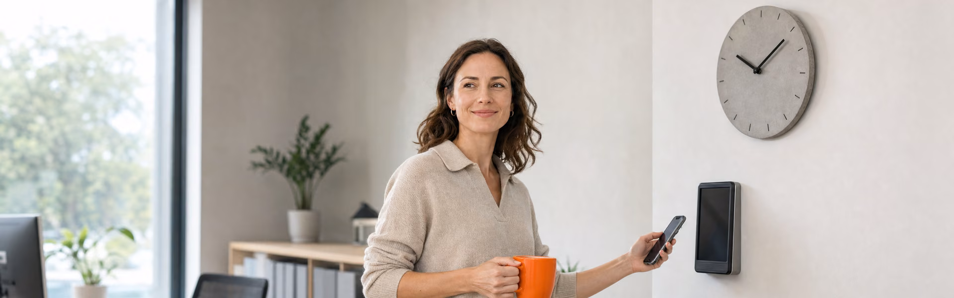 Mitarbeiterin mit Kaffeetasse stempelt sich im hellen Büro per Smartphone am Zeiterfassungsterminal ein, daneben eine Wanduhr